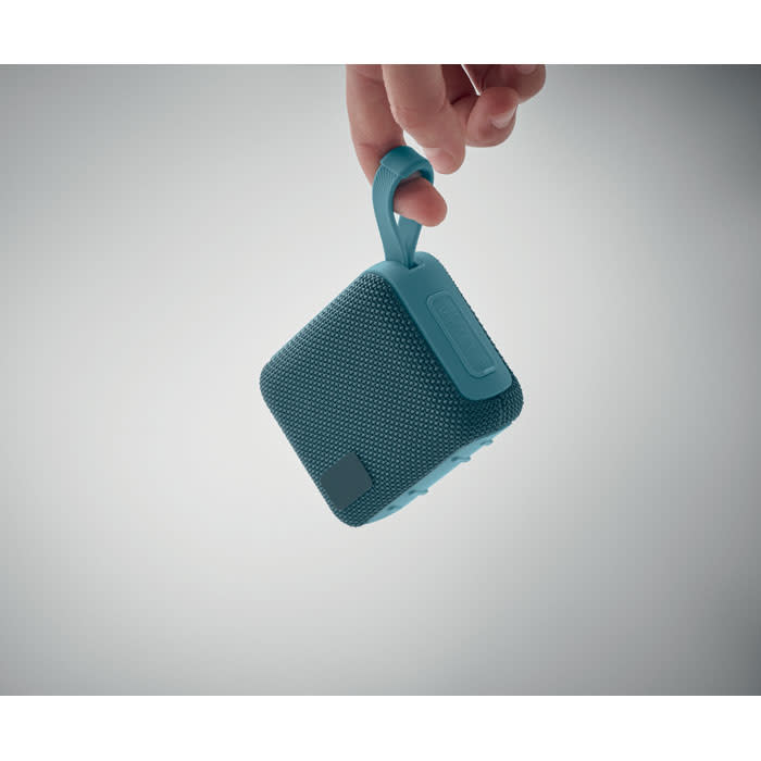 A portable speaker in teal, featuring a textured exterior and a carrying loop, held by a hand.