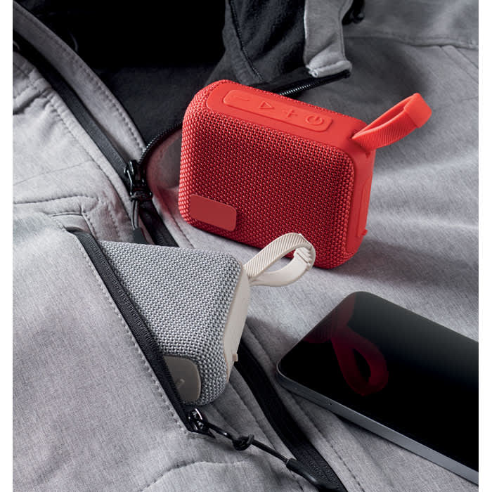 A portable speaker in red and grey sits in a zippered pocket alongside a smartphone. The speaker has a branding logo.
