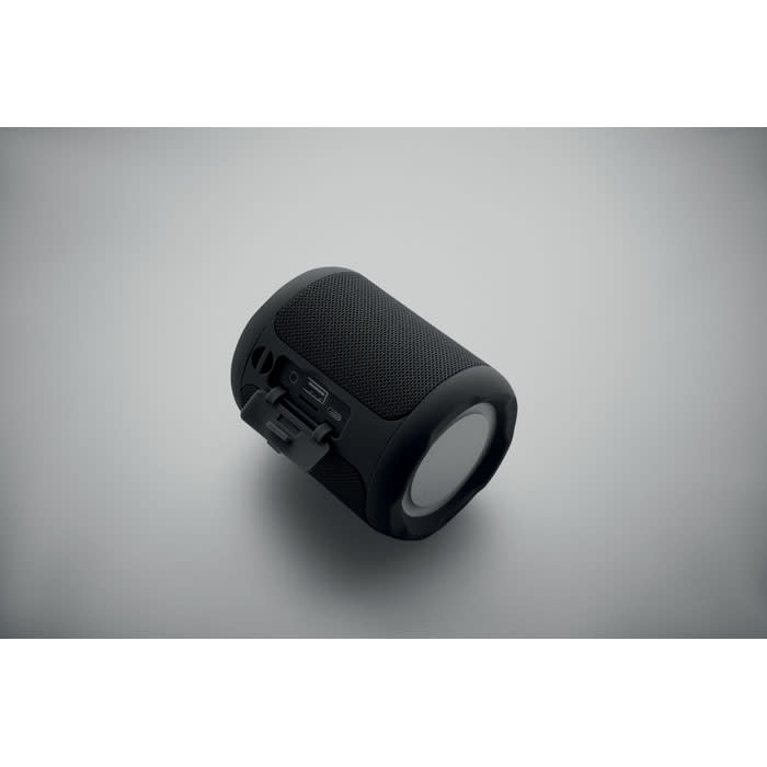 A portable speaker in black with a textured surface and a round speaker grille, featuring a built-in clip and ports.