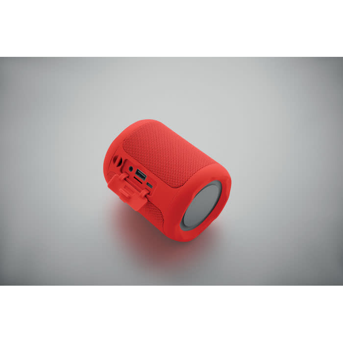 A portable speaker in bright red with a textured surface and a circular front. It has a logo and various ports on the side.