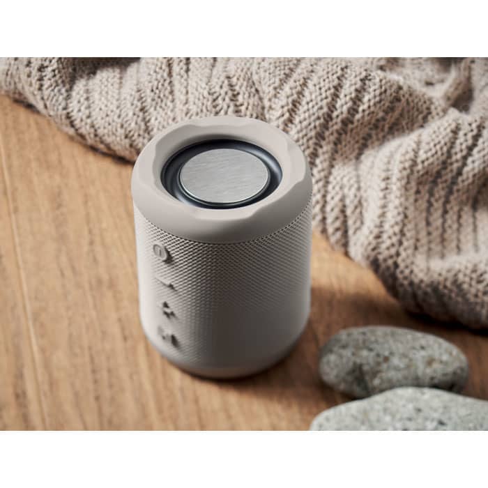 A portable speaker in light grey with a textured exterior and a round top, placed on a wooden surface with pebbles.