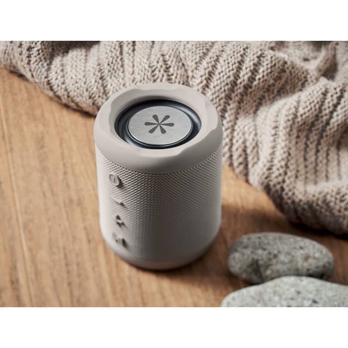 A portable speaker in a light grey colour with a textured surface, placed on a wooden surface beside smooth stones.