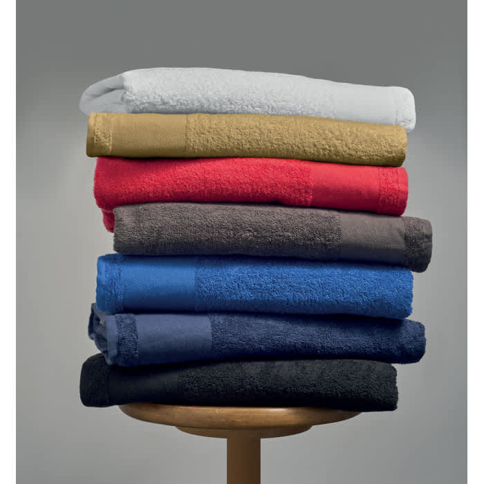 A stack of folded towels in various colours including white, gold, red, grey, blue, and black, displayed on a wooden stool.