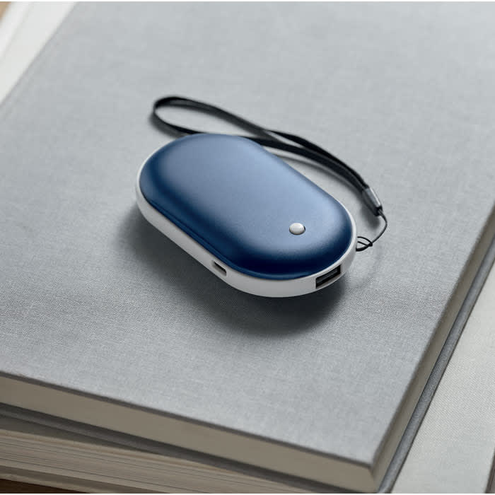A navy blue portable charger with a strap, positioned on a grey notebook. The charger has a small button on the front.