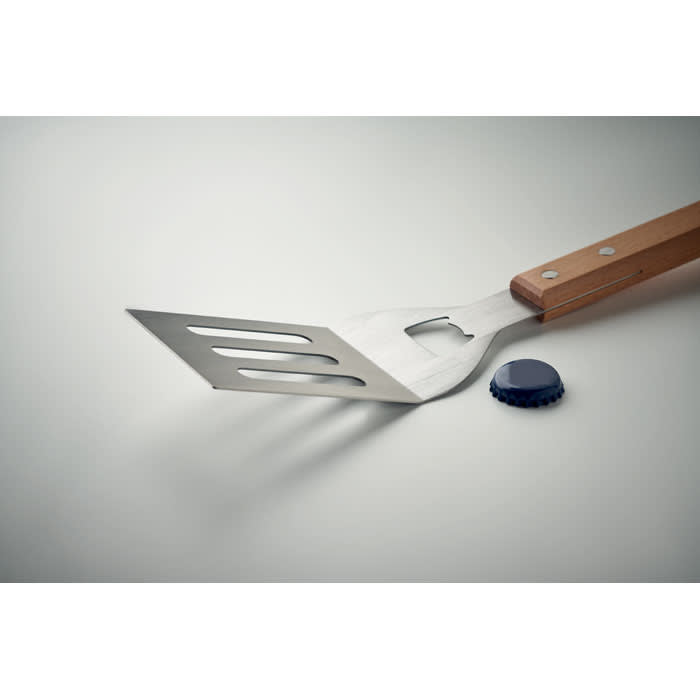 A stainless steel spatula with a wooden handle sits beside a blue bottle cap on a light surface.