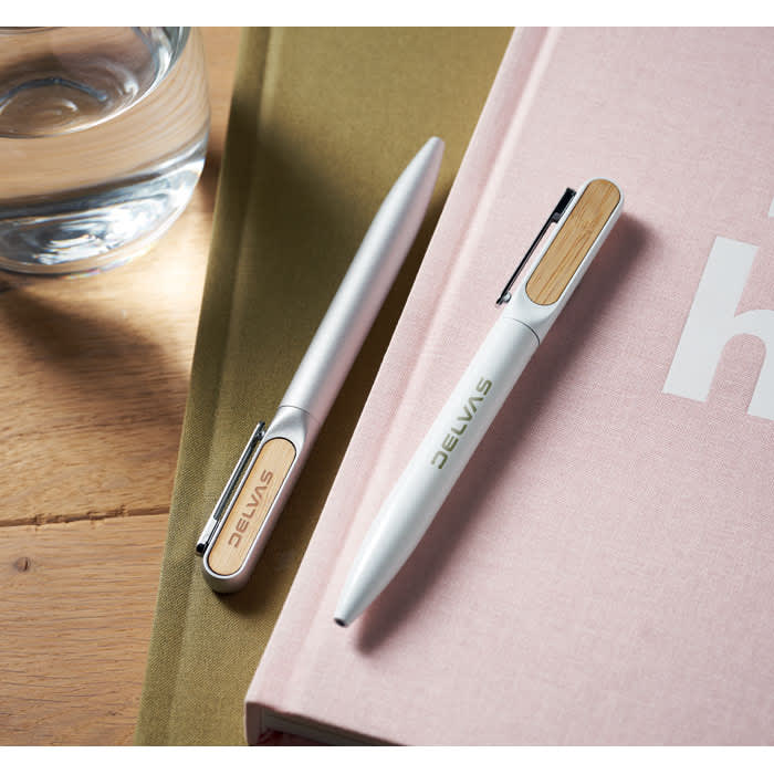 A set of two pens, one silver and one white with a wooden clip, resting on a pink book alongside a glass of water.