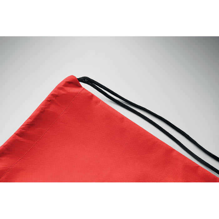 A red drawstring bag with black cords, featuring a smooth texture and angular shape. It has a branding logo on it.