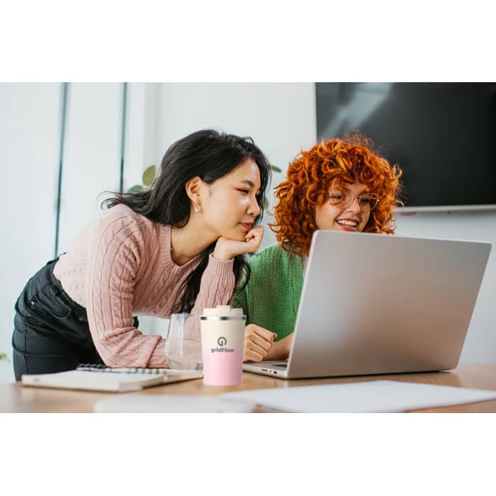 Two young women are looking at a laptop, sharing a moment together. One has dark hair, the other has curly red hair. A pink drink cup is on the table.