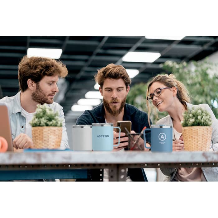 A drink bottle in muted grey sits on a table among three people chatting, with greenery and mugs visible in the background.