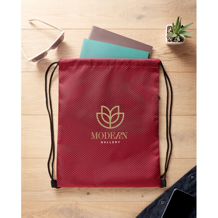 A red drawstring bag with a decorative logo, alongside sunglasses, notebook, and a small plant on a wooden surface.