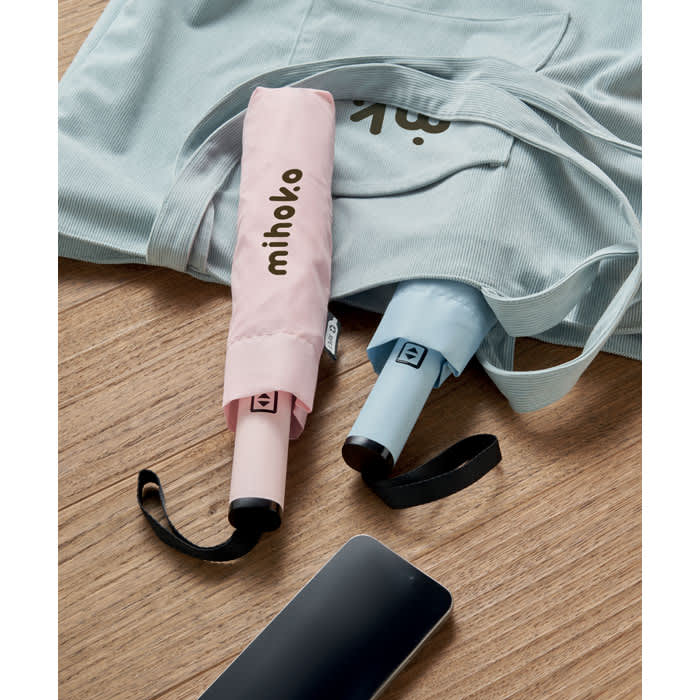 A set of compact umbrellas in pink and blue, resting on a wooden surface alongside a light blue tote bag.