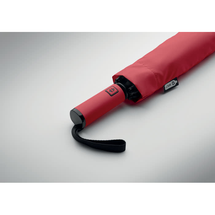 A red umbrella with a black handle and a strap, partially opened on a light background.