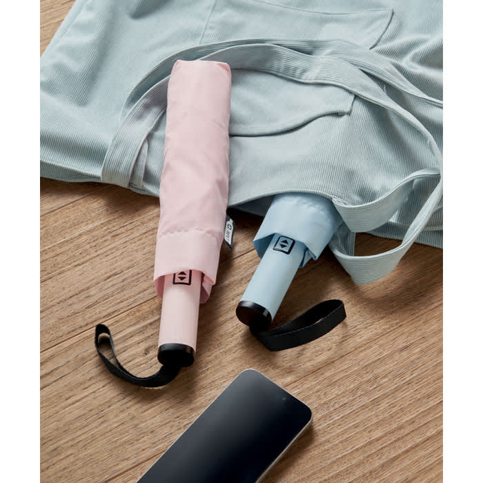 A pair of compact umbrellas in pink and blue resting on a wooden surface beside a bag and a phone.