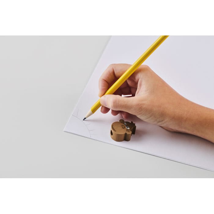 A hand holding a yellow pencil marks a corner on a sheet of white paper, with a brown bear-shaped sharpener nearby.