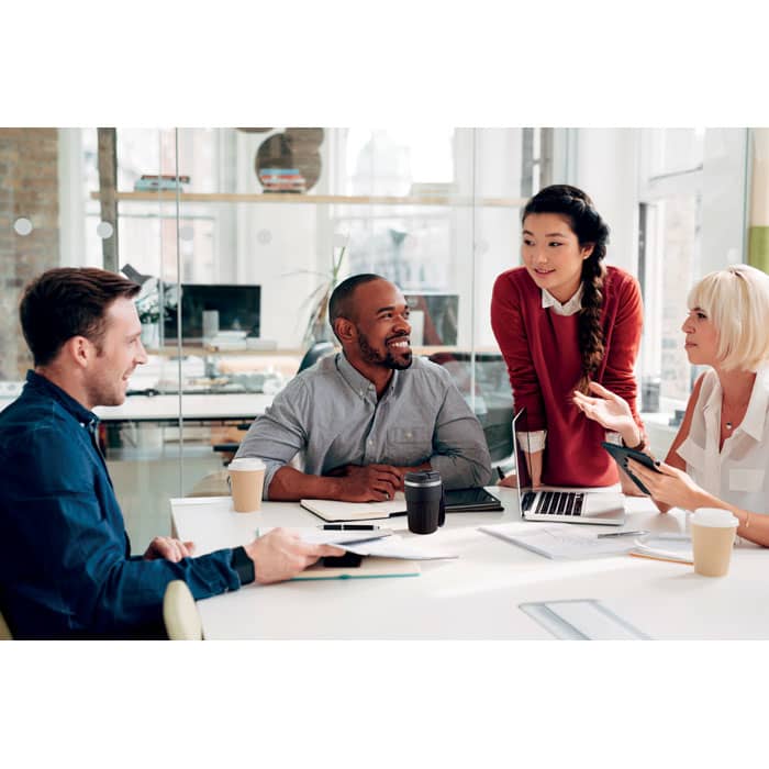 A group of four people engaged in discussion at a table with laptops and drinks in a modern office setting.