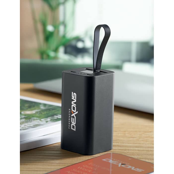 A portable black power bank featuring a strap and branding, sitting on a wooden table next to a notebook and card.