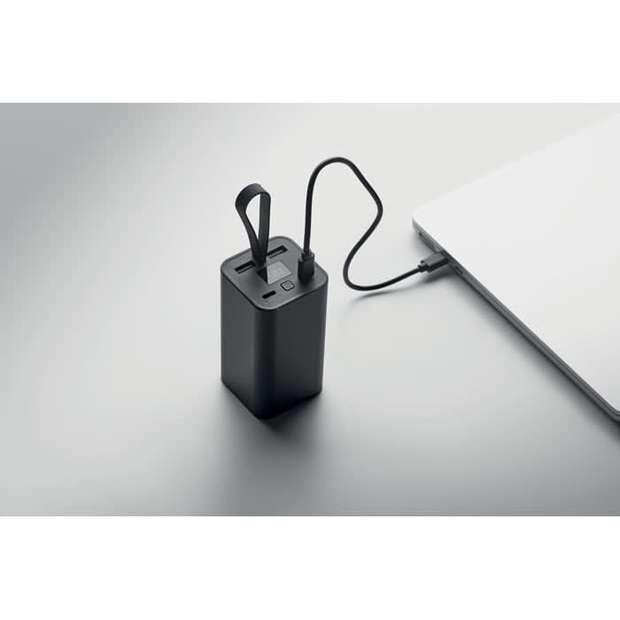 A power bank in black, with a charging cable connected to a laptop, on a light grey surface.