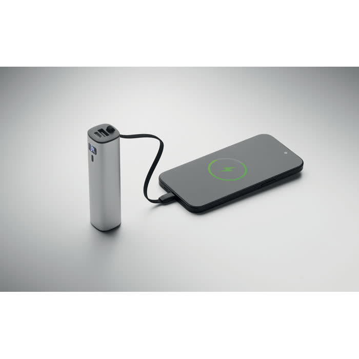 A silver power bank with a black charging cable connected to a smartphone on a wireless charging pad.