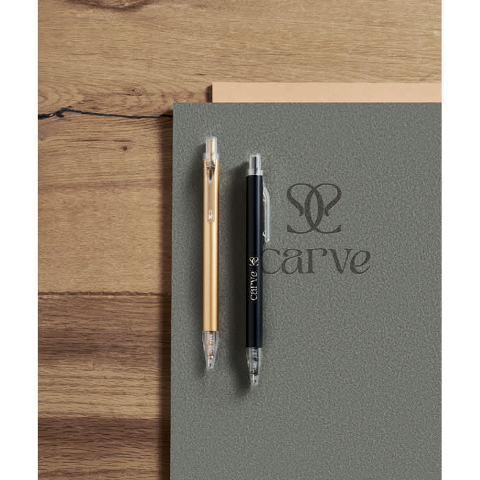 A notebook in grey with a logo, accompanied by a gold and a black pen on a wooden surface.