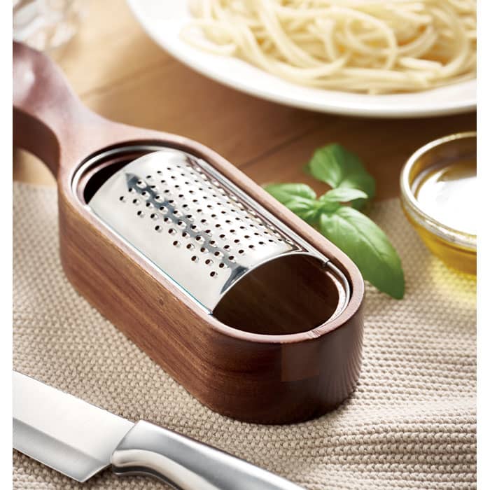 A grater with a wooden handle and stainless steel grating surface, placed beside pasta and basil leaves.