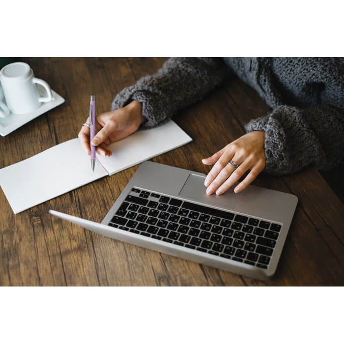 A desk with a laptop, a person writing on paper with a pen, and a coffee cup in the background.