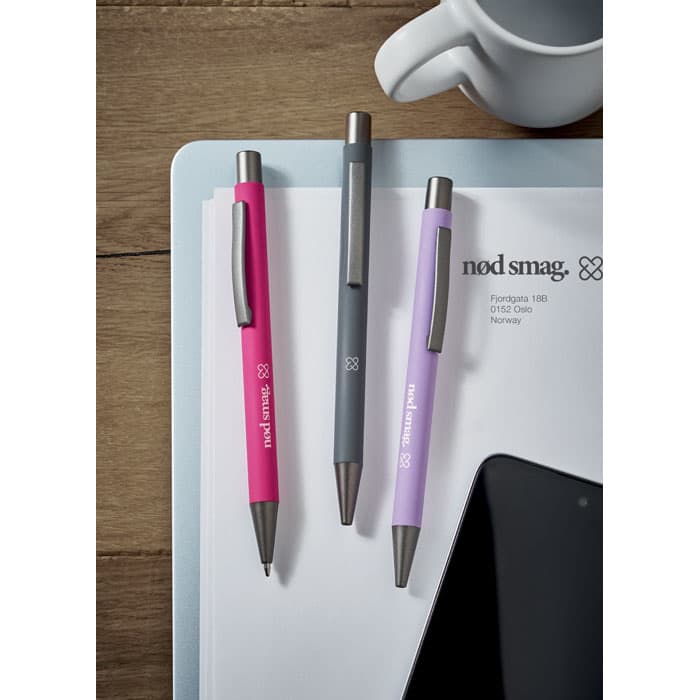 A collection of three pens in pink, grey, and purple, resting on a desk along with paper and a cup.