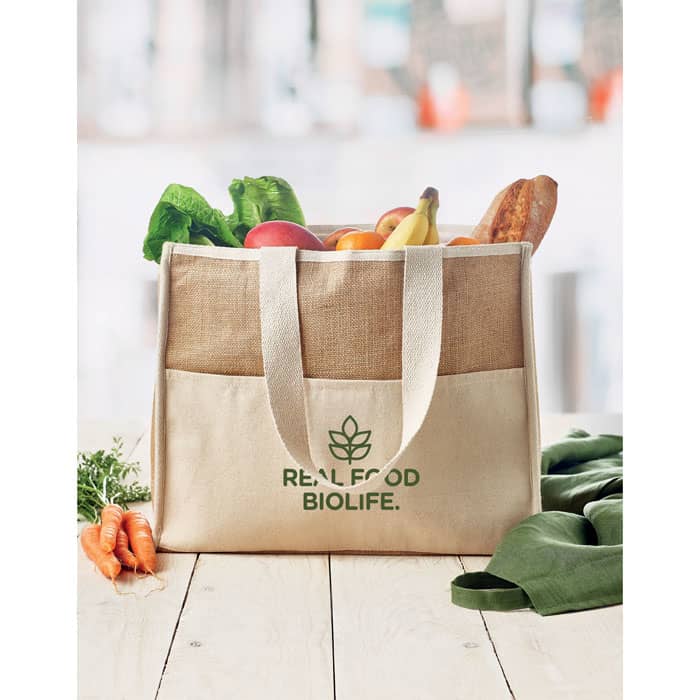 A tote bag in natural canvas with a logo, filled with fresh fruits and vegetables, featuring green and brown tones.