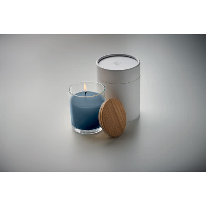 A candle in a glass holder, with a blue wax and wooden lid, next to a white cylindrical container.