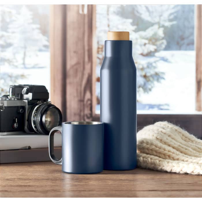 A drink bottle and mug in navy blue, resting on a wooden surface with a camera and a knitted item nearby.