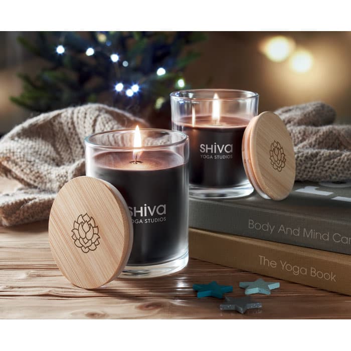 Two black candles in glass jars with wooden lids, featuring a logo, placed on a stack of books and a soft blanket.