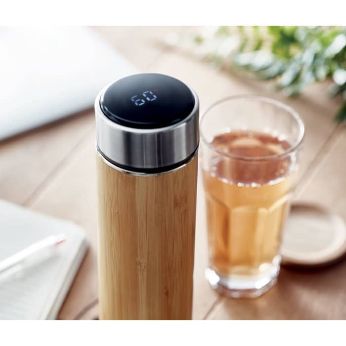 A drink bottle with a bamboo body and a stainless steel lid, alongside a glass of tea. It has a logo on the lid.