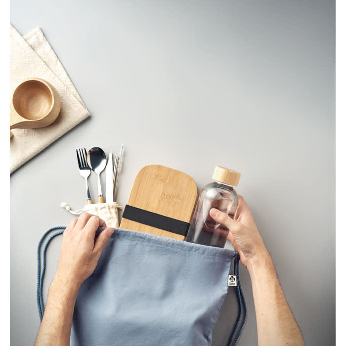 A drink bottle and bamboo lunch box are being placed into a blue fabric bag, surrounded by utensils and a wooden bowl.