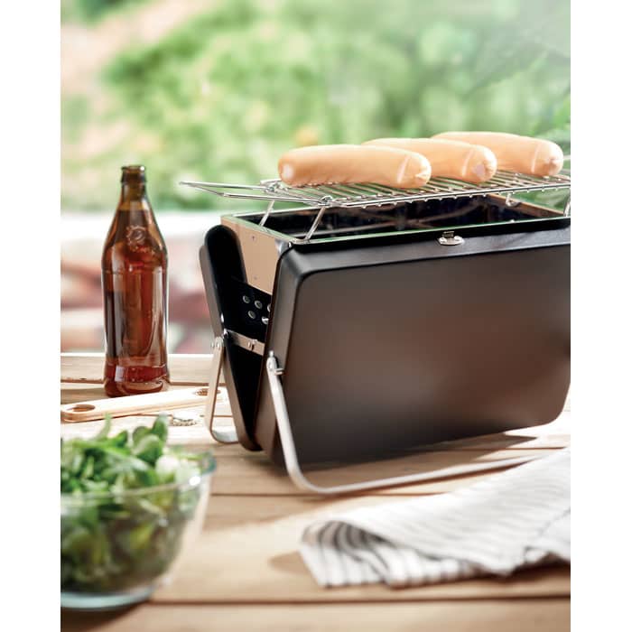 A portable black BBQ grill with sausages on the rack, accompanied by a drink bottle and a bowl of salad.
