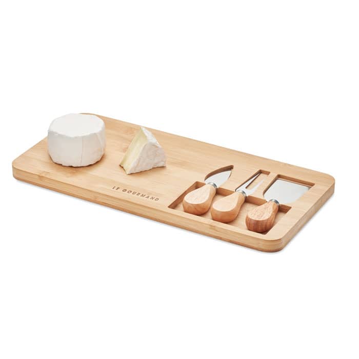 A cheese board made of bamboo, featuring a round cheese, a wedge of cheese, and three wooden utensils.