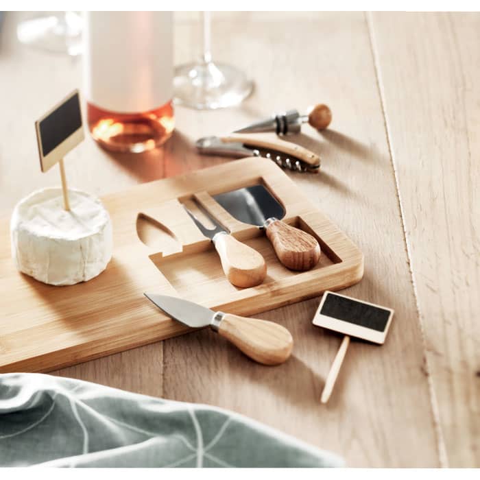 A wooden cheese board with various cheese knives, a small wooden scoop, and chalkboard labels on a wooden table.