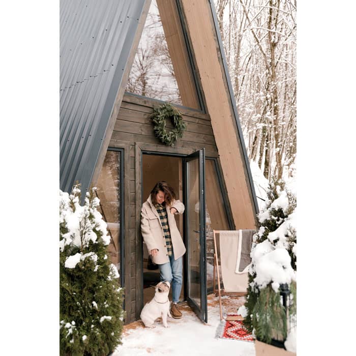 A woman stands outside an A-frame cabin, surrounded by snow, with a dog at her feet and evergreen decorations.