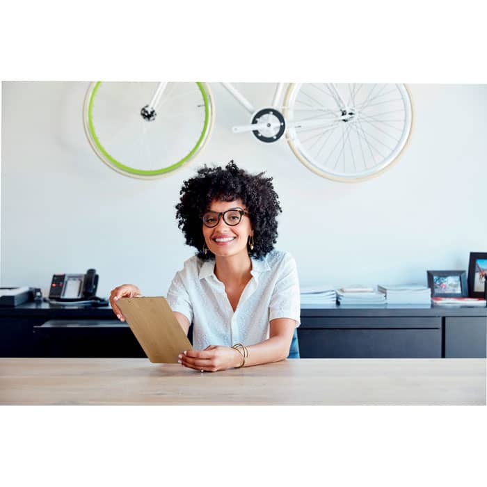 A woman with curly hair and glasses smiles while holding a clipboard in a modern office setting.