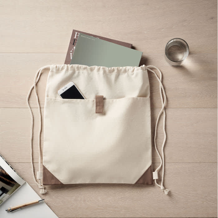 A cream and brown drawstring bag with a front pocket, containing a phone and some notebooks on a wooden surface.