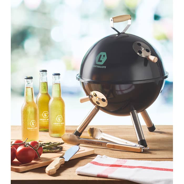 A black spherical barbecue with wooden handles, accompanied by three bottles, a knife, and red tomatoes on a table.