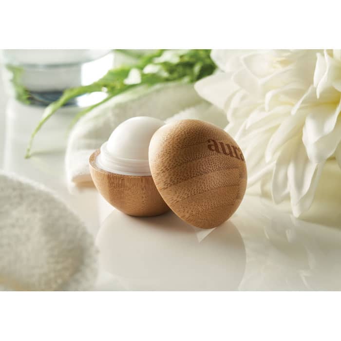 A bamboo container holding a white balm, with soft towels and flowers in the background, creating a calm aesthetic.