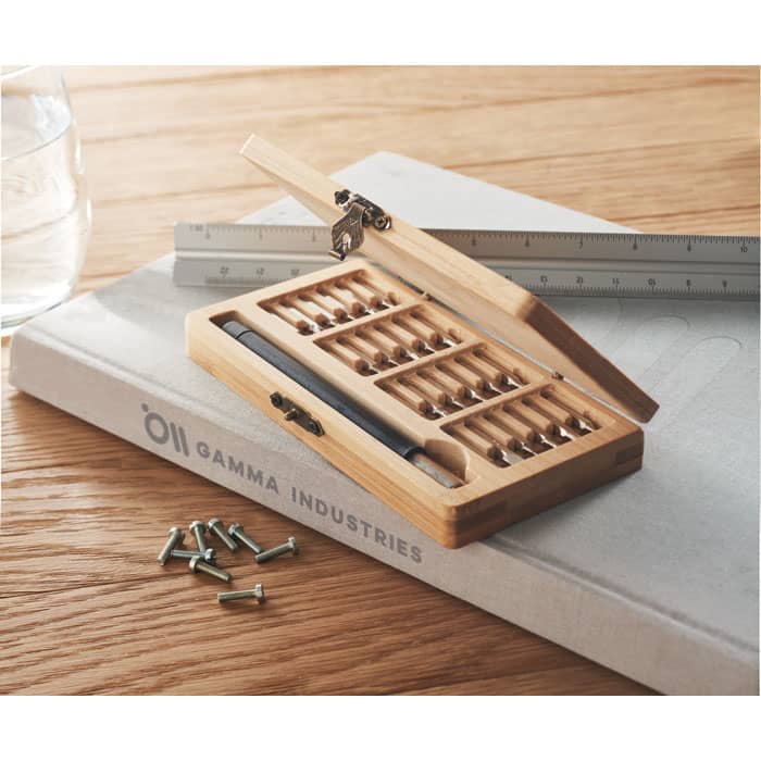 A wooden box containing various screws and a pen, placed on a wooden surface with a ruler and a notebook.