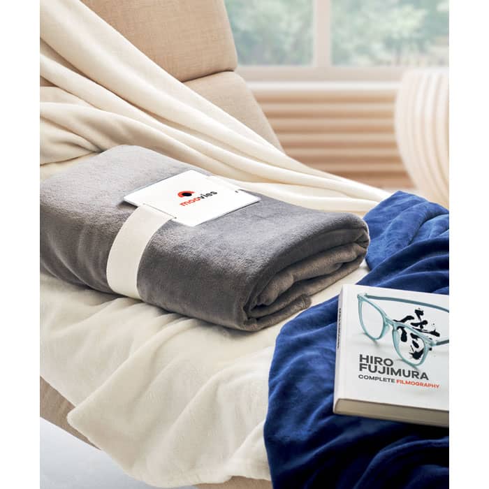 A soft grey blanket neatly rolled with a tag, placed on a beige chair alongside a book and a navy blue item.