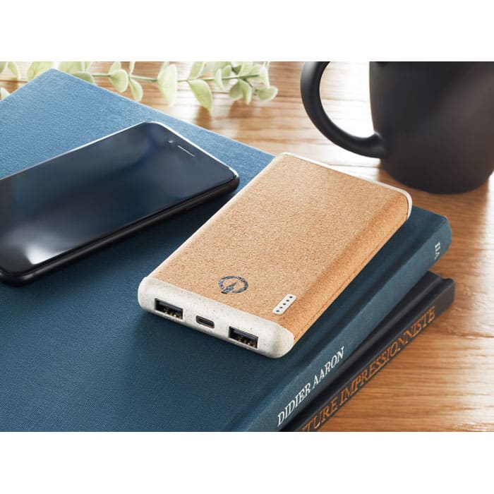 A power bank with a cork casing rests on books beside a smartphone and a coffee mug, on a wooden surface.