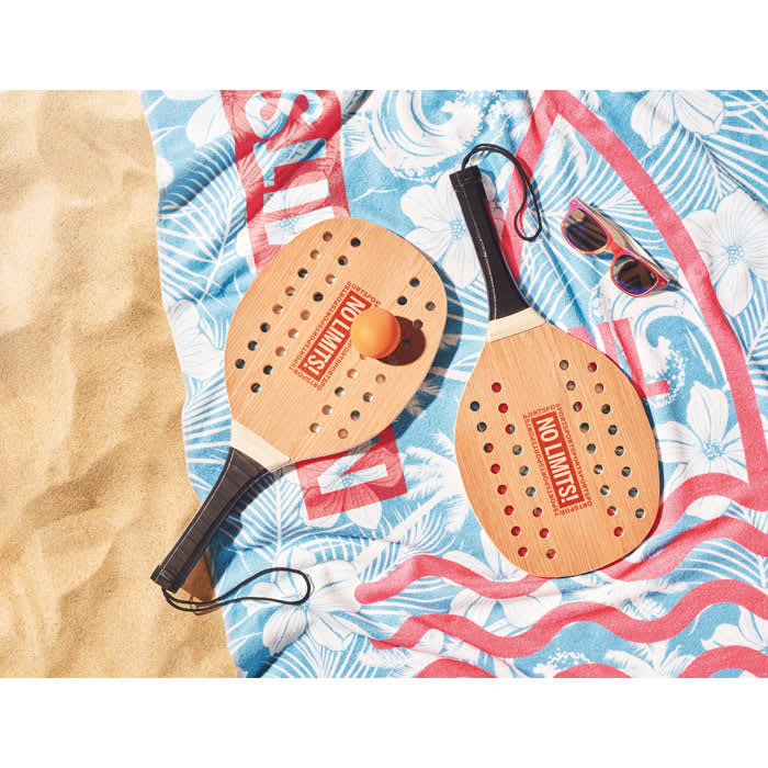 Two wooden beach paddles with a ball, resting on a patterned towel, alongside a pair of sunglasses.
