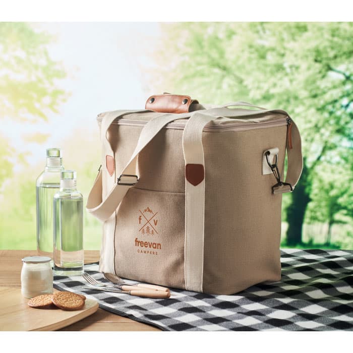 A drink bottle cooler in beige with leather handles, next to glass bottles, cookies, and a checkered cloth.