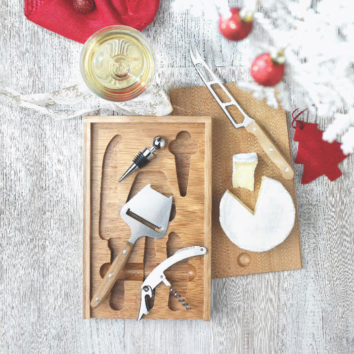 A wooden cheese board with cheese, a knife, corkscrew, and serving tools, accompanied by a glass of white wine.