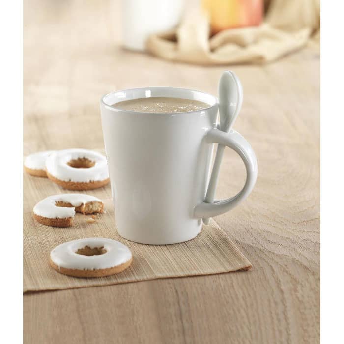 A white ceramic mug with a spoon attached, accompanied by circular cookies, on a wooden surface.