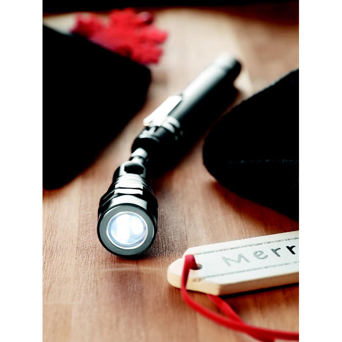 A black flashlight lies on a wooden surface, surrounded by soft fabric, with a festive tag nearby.
