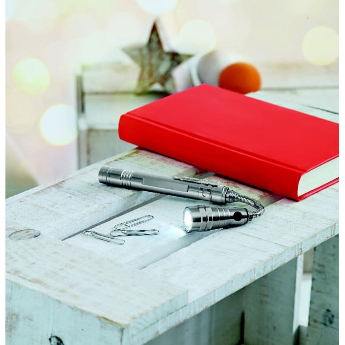 A silver flashlight and paper clips on a wooden surface beside a red notebook, with soft bokeh in the background.