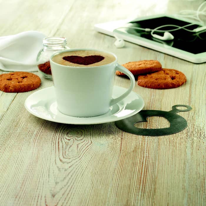 A white coffee cup with a heart-shaped design on top, alongside cookies and a tablet on a wooden table.